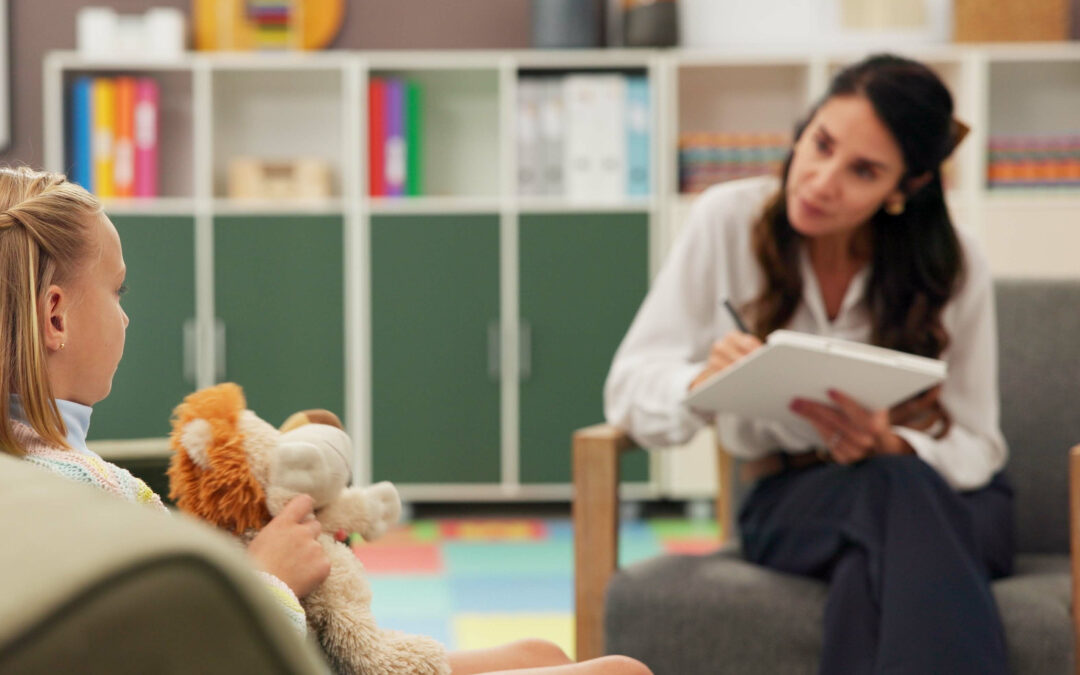 A young child holding a stuffed animal during an ADHD quick assessment session with a child psychologist taking notes in a brightly colored clinic room.