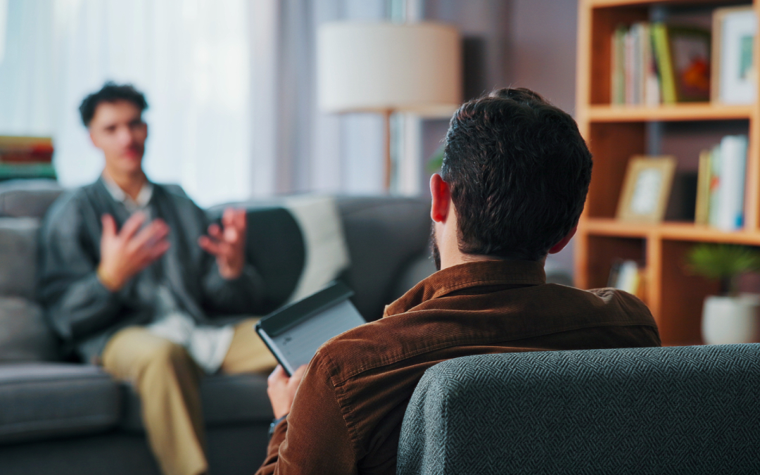 A therapist sitting in an armchair with a digital tablet listening to a client speak on a sofa.