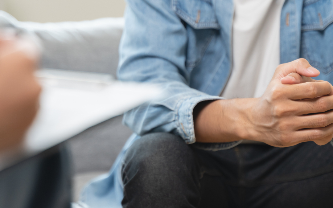 Close-up of a client in a denim jacket sitting on a couch with clasped hands, speaking with a psychologist holding a clipboard in the foreground.